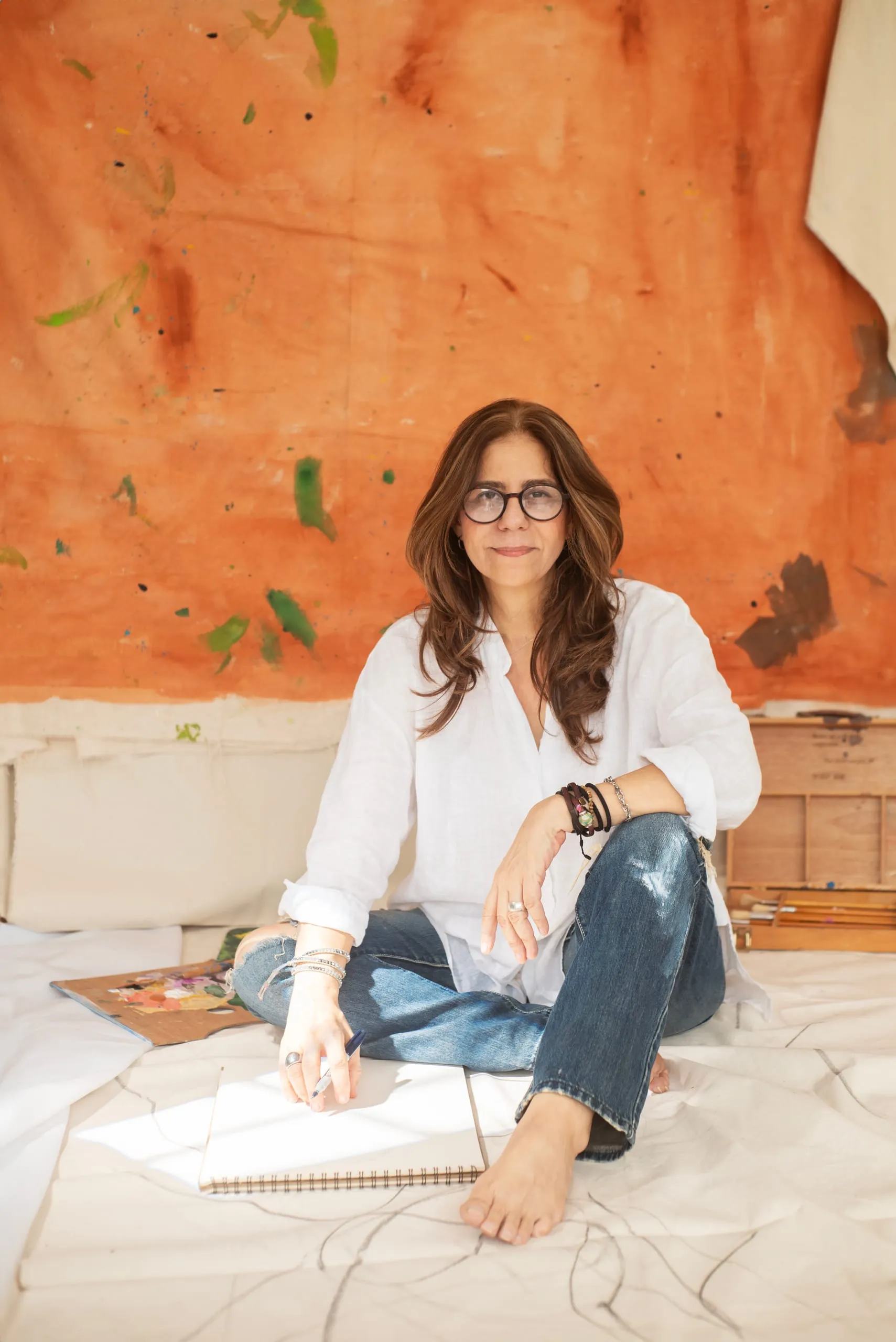 Elsa Basil seated in her studio in front of a paint-covered orange wall